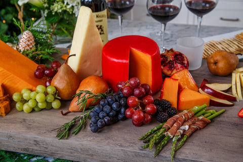 Assorted cheeses, fruits, and vegetables on a wooden board with wine glasses in the background.